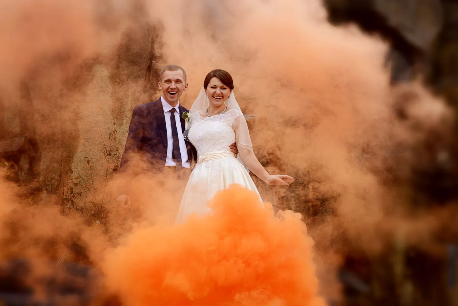 Happy bride and groom surrounded by vibrant orange smoke outdoors, exuding joy and celebration.